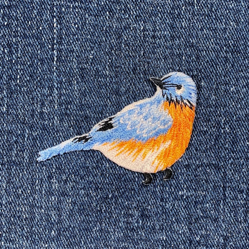 Pu&ograve; includere: Toppa ricamata di un uccello azzurro con dettagli blu, arancioni e neri. L'uccello &egrave; cucito su uno sfondo di denim. La toppa &egrave; un oggetto decorativo, adatto per abbigliamento o accessori.