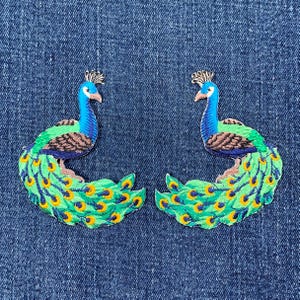 May include: Two embroidered peacock patches. Each patch has a blue head and neck, green body, and a fan of green and yellow tail feathers. The patches are on a blue denim background.