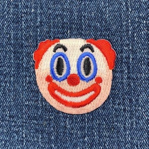 May include: Embroidered patch of a clown face with red hair, blue eyes, and a red smile. The patch is on a blue denim background.