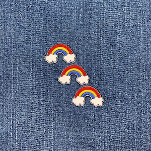 May include: Three embroidered rainbow patches with red, yellow, and blue arches and white cloud accents are arranged on a blue denim background. The patches are small and colorful, suitable for clothing or accessories.