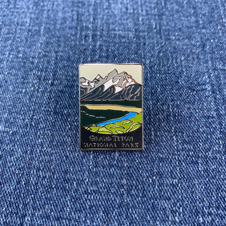 May include: A rectangular enamel pin featuring a landscape of mountains, a river, and a valley. The pin has a black border and the text "GRAND TETON NATIONAL PARK" at the bottom. The mountains are gray and white.
