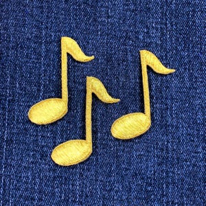 May include: Three embroidered gold musical note patches on a blue denim background. The notes are detailed with a textured appearance, showcasing the treble clef shape. These patches are suitable for fabric crafts and music-themed projects.