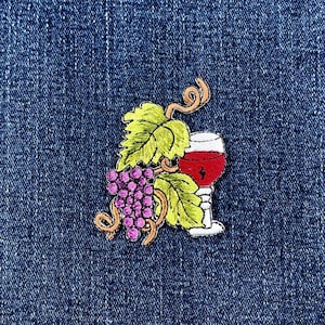May include: Embroidered patch featuring a wine glass filled with red liquid, a cluster of purple grapes, and green leaves. The patch has a white border and is set against a blue denim background. The design includes brown vines.