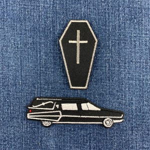 May include: Two embroidered patches featuring a black coffin with a white cross outline and a black hearse vehicle with white detailing. The patches are stitched with black, white, and a small red accent on the rear of the hearse vehicle.