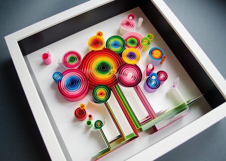 May include: A colorful paper quilling art piece in a white shadow box frame. The artwork features concentric circles in a rainbow of colors, with additional rolled paper elements and linear designs. The background is white, and the frame is white.