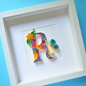 May include: A white frame with a colorful paper quilling art piece of the letter R. The letter is decorated with flowers and a butterfly.