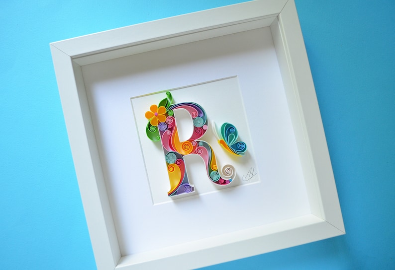May include: A white frame with a colorful paper quilling art piece of the letter R. The letter is decorated with flowers and a butterfly.