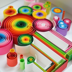 May include: A colorful paper quilling art piece featuring concentric circles and rolled paper shapes in a variety of vibrant colors, including red, orange, yellow, green, blue, and pink. The artwork is arranged in a white tray.