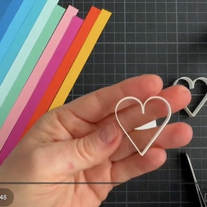 May include: A white heart-shaped paper quilling form held in the center, with a second identical heart form nearby. Beside these are strips of paper in a rainbow gradient, a small bottle of white glue, a white tool, and a circular logo sticker.
