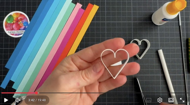 May include: A white heart-shaped paper quilling form held in the center, with a second identical heart form nearby. Beside these are strips of paper in a rainbow gradient, a small bottle of white glue, a white tool, and a circular logo sticker.