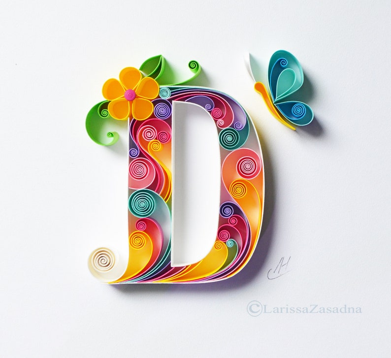 May include: A colorful paper quilling art piece depicting the letter D with a flower and butterfly design. The letter is made of curled paper strips in shades of pink, yellow, blue, green, and purple.