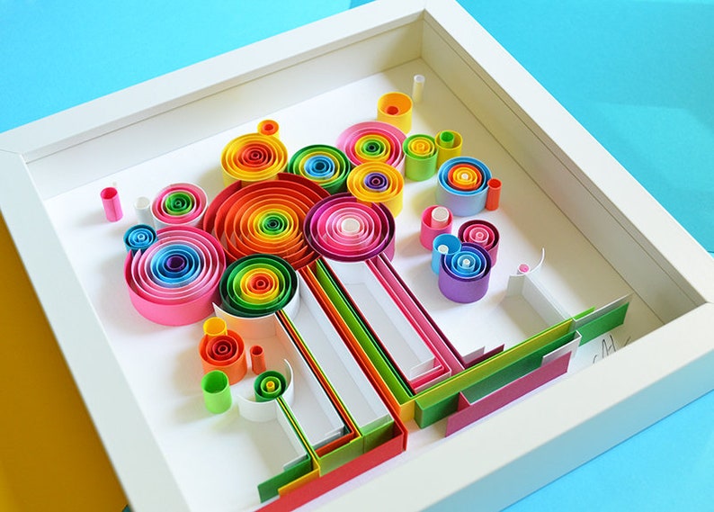 May include: A colorful paper quilling art piece in a white shadow box frame. The artwork features vibrant, rolled paper elements in various sizes and colors, including pink, orange, green, and blue, arranged in a whimsical design.