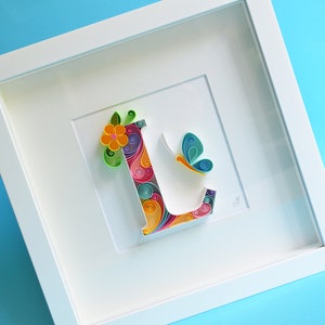 May include: A white frame with a colorful paper quilling art piece of the letter L. The letter is decorated with a flower and a butterfly.