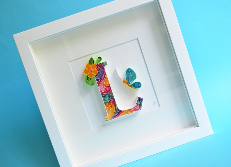 May include: A white frame with a colorful paper quilling art piece of the letter L. The letter is decorated with a flower and a butterfly.