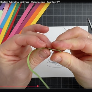 May include: A light green paper strip being folded by fingers for a quilling project. The scene includes a white card with a Christmas tree outline, various colorful paper strips, a white glue bottle, and metal tweezers on a dark grid mat.