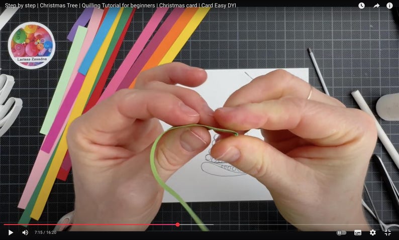 May include: A light green paper strip being folded by fingers for a quilling project. The scene includes a white card with a Christmas tree outline, various colorful paper strips, a white glue bottle, and metal tweezers on a dark grid mat.