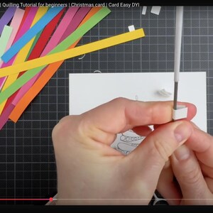 May include: A quilling tool with a white handle is being used to roll a small white paper strip. A pile of colorful paper strips in shades of yellow, orange, red, pink, purple, blue, and green lies nearby on a dark grid mat next to a white card.
