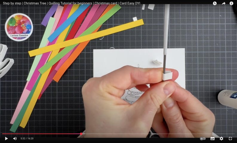 May include: A quilling tool with a white handle is being used to roll a small white paper strip. A pile of colorful paper strips in shades of yellow, orange, red, pink, purple, blue, and green lies nearby on a dark grid mat next to a white card.
