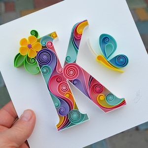 May include: A colorful paper quilling art piece featuring the letter K, a yellow flower, and a blue butterfly. The letter K is made of swirls of pink, blue, green, and yellow paper.