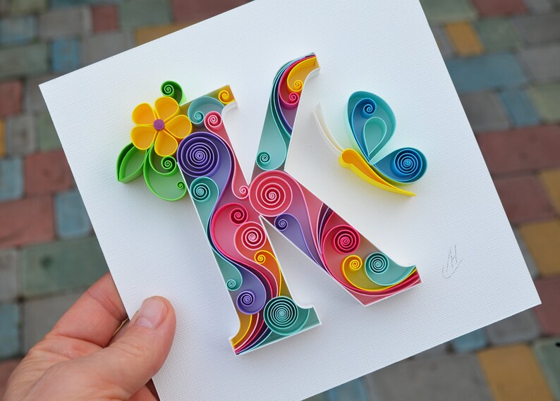 May include: A colorful paper quilling art piece featuring the letter K, a yellow flower, and a blue butterfly. The letter K is made of swirls of pink, blue, green, and yellow paper.