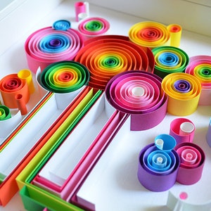 May include: A vibrant, colorful paper quilling art piece. The artwork features concentric circles and tubes in a rainbow of hues, including red, orange, yellow, green, blue, and purple. The design is arranged in a white tray, creating a visually appealing display.