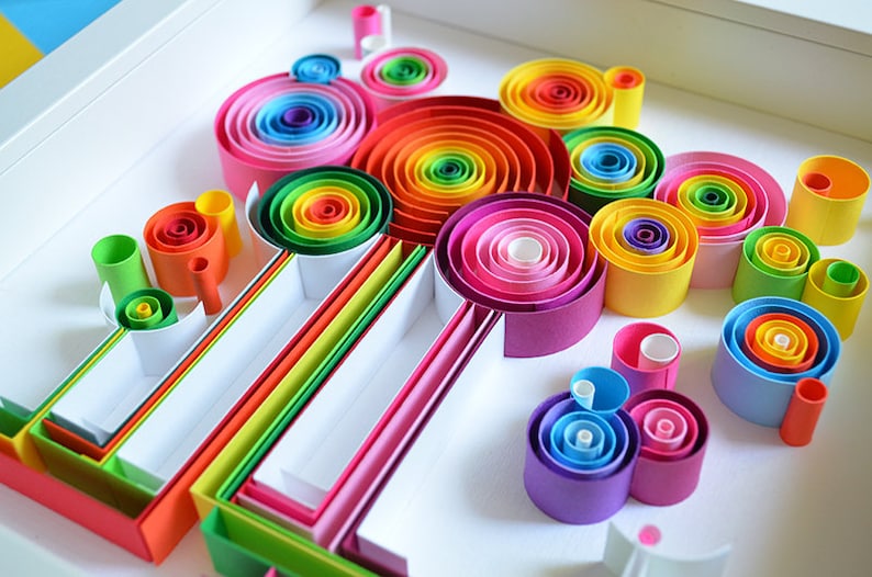 May include: A vibrant, colorful paper quilling art piece. The artwork features concentric circles and tubes in a rainbow of hues, including red, orange, yellow, green, blue, and purple. The design is arranged in a white tray, creating a visually appealing display.