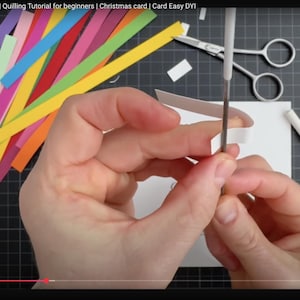 May include: A quilling tool with a white handle and metal tip is used to coil a white paper strip. Nearby are colorful paper strips in yellow, orange, red, pink, purple, and green, along with silver scissors and a white card on a black grid mat.