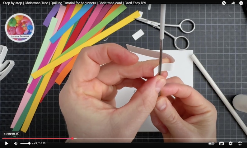 May include: A quilling tool with a white handle and metal tip is used to coil a white paper strip. Nearby are colorful paper strips in yellow, orange, red, pink, purple, and green, along with silver scissors and a white card on a black grid mat.
