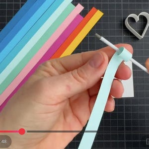 May include: A light blue paper strip being coiled around a white quilling tool. Beside the tool are several colorful paper strips in shades of blue, green, pink, orange, and yellow, along with a white heart-shaped mold and a circular logo.
