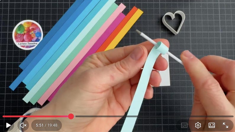 May include: A light blue paper strip being coiled around a white quilling tool. Beside the tool are several colorful paper strips in shades of blue, green, pink, orange, and yellow, along with a white heart-shaped mold and a circular logo.