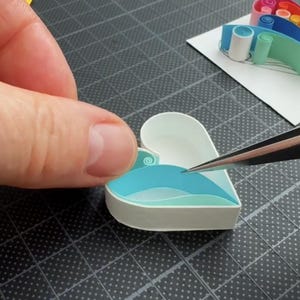 May include: A heart-shaped paper quilling project featuring white outer edges and layered light blue and teal paper strips inside. A separate rainbow-colored paper quilling element sits on a white square nearby, while metal tweezers are used.