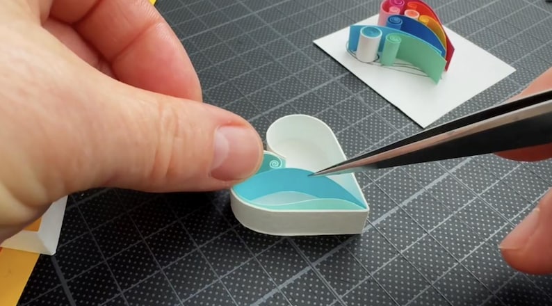 May include: A heart-shaped paper quilling project featuring white outer edges and layered light blue and teal paper strips inside. A separate rainbow-colored paper quilling element sits on a white square nearby, while metal tweezers are used.