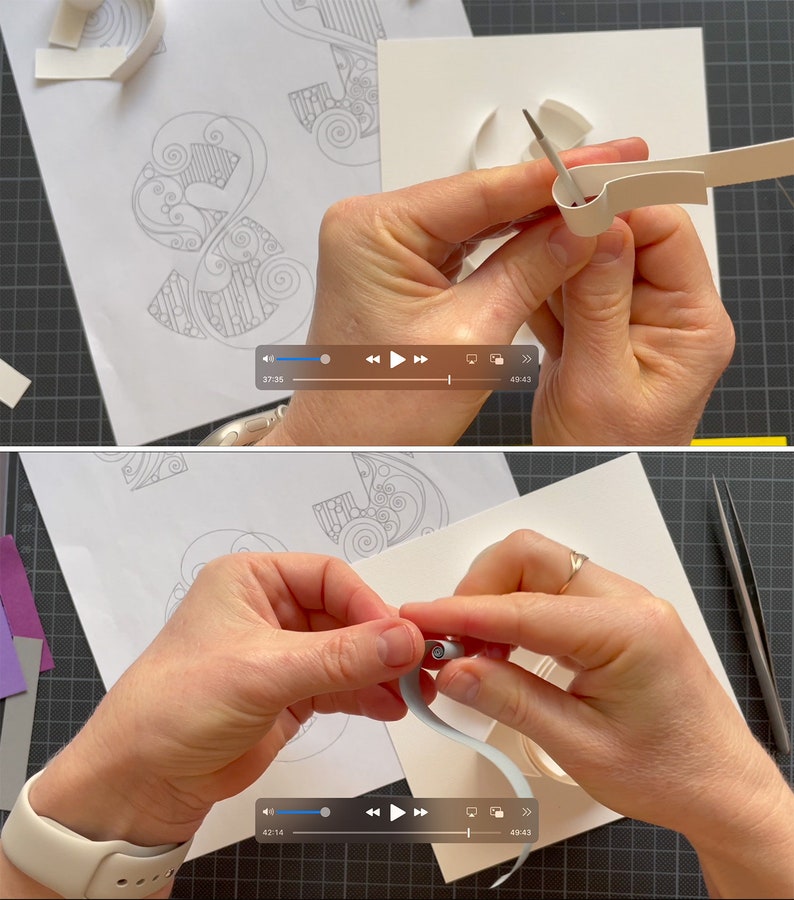 May include: Close-up of a person's hands carefully folding a white paper strip to create a three-dimensional design. The paper strip is being folded around a small, round object, creating a curved shape. The person is using their fingers to hold the paper in place as they fold it.