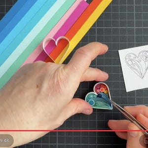 May include: A paper quilling project featuring a heart shape filled with colorful coiled paper strips in shades of blue, green, red, and orange. A white square card with a black line drawing of a heart and a set of rainbow-colored paper strips are visible.