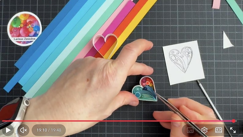 May include: A paper quilling project featuring a heart shape filled with colorful coiled paper strips in shades of blue, green, red, and orange. A white square card with a black line drawing of a heart and a set of rainbow-colored paper strips are visible.