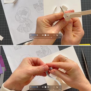 K&ouml;nnte beinhalten: Ein Papier-Quilling-Projekt mit wei&szlig;en Papierstreifen, die mit einem Metallwerkzeug geformt werden. Der Prozess umfasst das Rollen und Bearbeiten d&uuml;nner Papierstreifen auf einer wei&szlig;en Oberfl&auml;che mit gedruckten Mustern, was die Kunst des Papierfiligrans zeigt.