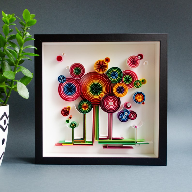 May include: A framed paper quilling art piece featuring colorful, concentric circles in shades of red, orange, yellow, green, and blue. The artwork is set against a white background within a black frame, creating a vibrant and eye-catching display.
