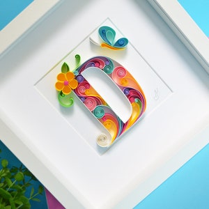 May include: A white frame with a colorful paper quilled letter D, a yellow flower, and a blue butterfly. The letter D is made of paper that is rolled into spirals and then arranged in a pattern.