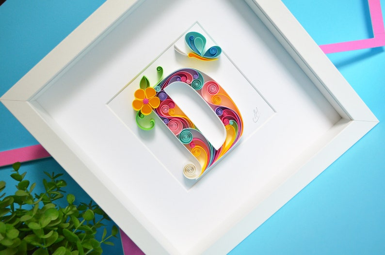 May include: A white frame with a colorful paper quilled letter D, a yellow flower, and a blue butterfly. The letter D is made of paper that is rolled into spirals and then arranged in a pattern.