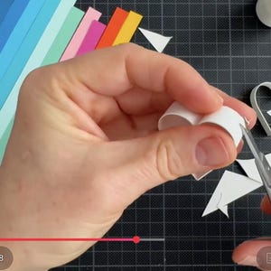 May include: A white paper strip being shaped into a heart using metal scissors. Beside the heart are colorful paper strips in shades of blue, green, pink, and orange, a white heart-shaped mold, a glue bottle, and a thin metal tool.