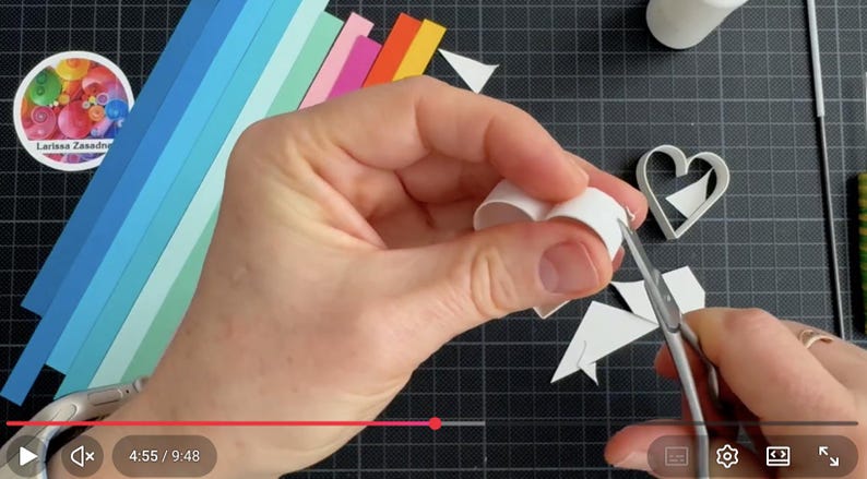 May include: A white paper strip being shaped into a heart using metal scissors. Beside the heart are colorful paper strips in shades of blue, green, pink, and orange, a white heart-shaped mold, a glue bottle, and a thin metal tool.