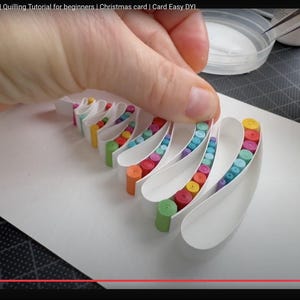 May include: A white paper card featuring a quilled Christmas tree design made of curved white paper strips filled with small, tightly rolled colorful paper coils in shades of green, blue, purple, red, orange, and yellow, arranged in a layered pattern.