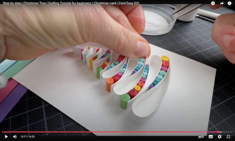 May include: A white paper card featuring a quilled Christmas tree design made of curved white paper strips filled with small, tightly rolled colorful paper coils in shades of green, blue, purple, red, orange, and yellow, arranged in a layered pattern.