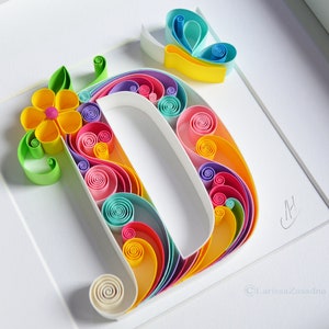May include: A colorful paper quilling art piece featuring the letter D with a flower and butterfly design. The letter is made of rolled paper strips in shades of pink, blue, yellow, green, and white. The flower is yellow with a pink center, and the butterfly is blue and yellow. The artwork is signed "CH" and "Larissa Zasadina".