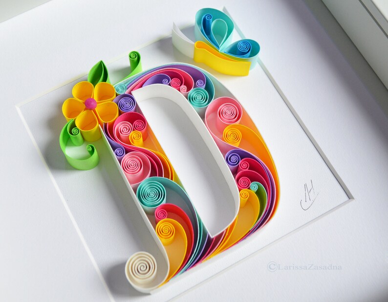 May include: A colorful paper quilling art piece featuring the letter D with a flower and butterfly design. The letter is made of rolled paper strips in shades of pink, blue, yellow, green, and white. The flower is yellow with a pink center, and the butterfly is blue and yellow. The artwork is signed "CH" and "Larissa Zasadina".