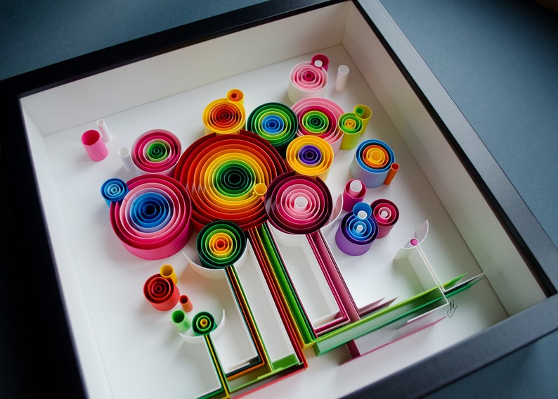 May include: A framed paper quilling art piece featuring a colorful abstract design. The artwork showcases concentric circles in a rainbow of colors, along with other rolled paper shapes, all set against a white background.