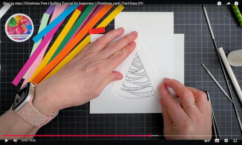 May include: A white card featuring a black line drawing of a Christmas tree with swirling patterns, surrounded by colorful paper strips in shades of pink, blue, green, orange, and yellow, along with crafting tools including tweezers and a white glue stick.
