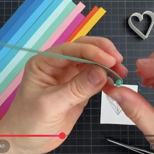May include: A light green paper strip being coiled by hand for quilling, with a stack of colorful paper strips in shades of blue, pink, orange, and yellow nearby. A white heart-shaped mold and a small square paper with a line drawing are present.