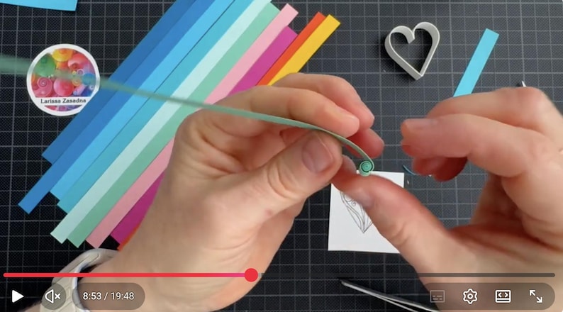 May include: A light green paper strip being coiled by hand for quilling, with a stack of colorful paper strips in shades of blue, pink, orange, and yellow nearby. A white heart-shaped mold and a small square paper with a line drawing are present.