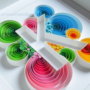 Op de afbeelding: Een wit frame met een kleurrijk papier-quilling kunstwerk in de vorm van de letter K. Het kunstwerk is gemaakt van concentrische cirkels van papier in tinten van groen, blauw, roze, oranje en geel.